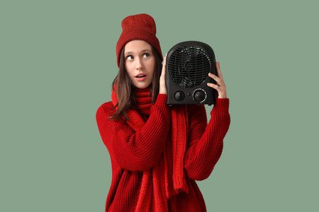 Young woman in winter clothes with electric fan heater on green backgroundの写真素材