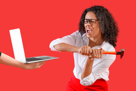 Angry African-American businesswoman with hammer breaking laptop on red backgroundの写真素材