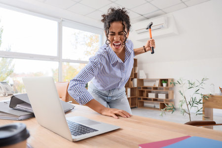 Angry African-American businesswoman with hammer breaking laptop in officeの写真素材