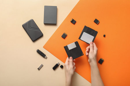 Hands holding floppy disks with modern digital storage devices on orange and beige background, this visual comparison highlights the evolution of computing and information storage over time