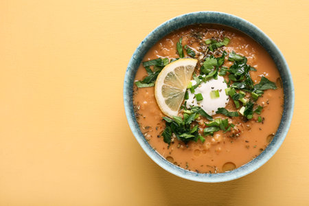 Bowl of tasty lentil soup with parsley and lemon on yellow background