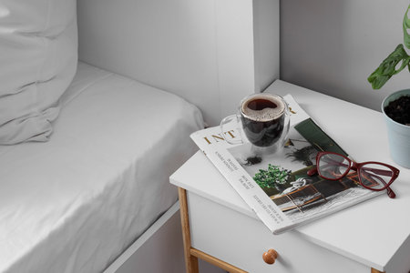 Morning routine and relaxation, coffee cup with magazine and reading glasses on a bedside table in a bedroom