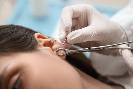 Master holding forceps and earring for ear piercing of young woman in studio, closeup