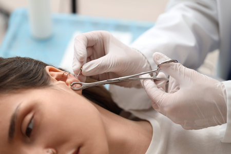Master holding forceps and earring for ear piercing of young woman in studio, closeup