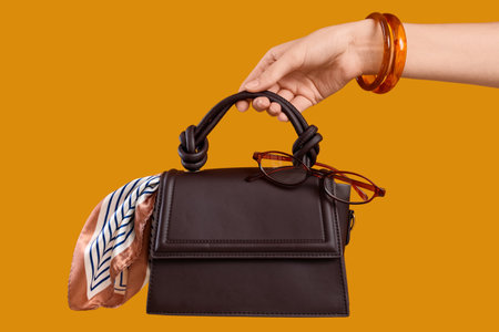 Fashion and luxury lifestyle, woman holding a dark brown handbag with a silk scarf and reading glasses on a solid mustard yellow background