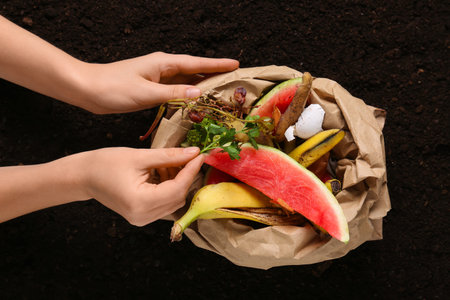 Female hand with paper bag of organic waste on soil background. Compost recycling concept