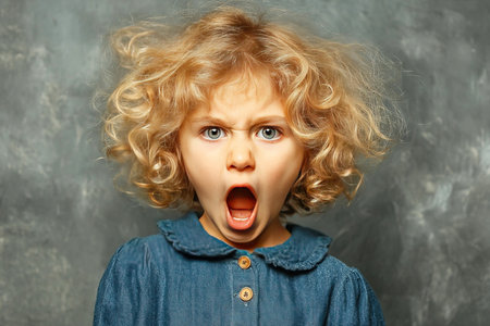 A young child with curly hair is expressing rage screaming with tears in their eyes. The background is simple and muted focusing on the child's emotional turmoil.の素材