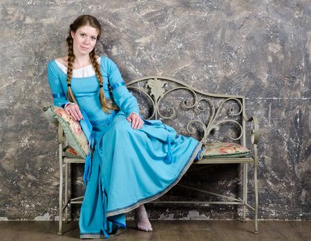 Pretty young woman in historical medieval blue sits on a banquette in studioの写真素材