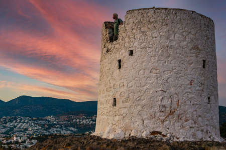 The ruins of an ancient mill, symbol of Bodrum, on a hill. Below is one of the districts of Bodrum Gumbet. Evening, colored sky. Man looking into the distance.のeditorial素材