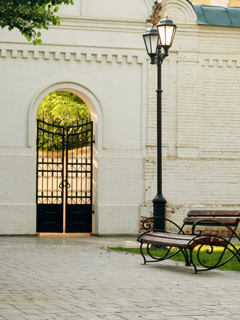 Streetlight and bench in city park. Shooting time - morning, spring. In the foreground a bench and a lamp. On a background - a white brick wall with an arch and gate. Vertical format. Outdoors. Without people. Color. Photo.の写真素材