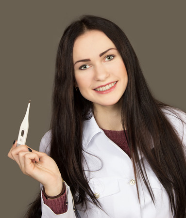 the young woman the doctor in a white scrub with the electronic thermometer in a hand on a gray backgroundの写真素材