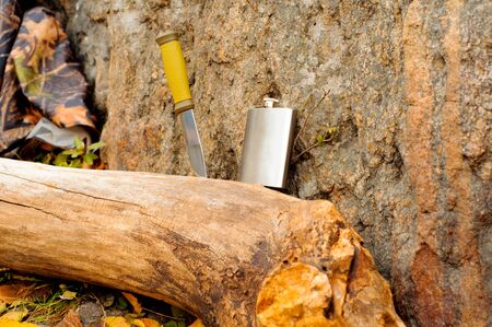 Metal flask and a knife on a log next to a raincoat of protective colorの写真素材