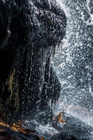 Close-up splashes water flying in different directions. Water flowing off cliff. Drops water falling on granite stones. Cobblestones overgrown with green moss. Yellow autumn fallen leaves. Outdoors.の写真素材