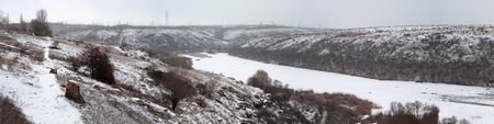 Winter picturesque panorama on banks of river. Winter beautiful landscape near small town. Beautiful panoramic view of hilly coastline to river in cloudy weather. Horizontal photo. Outdoors.の写真素材