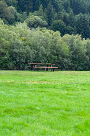 Wooden bench and table on green lawn near river among mountains and dense forestの写真素材