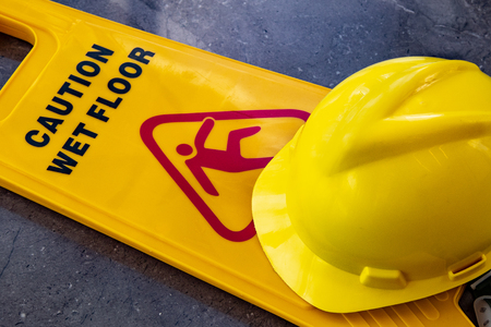 yellow plastic sign showing caution warning of wet floor with yellow protective helmet lying on floorの写真素材