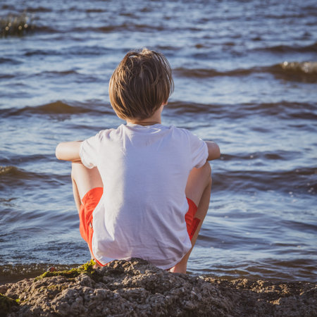 child sits on the seashore, boy looks into the sea at sunset, concept of summer vacation and...