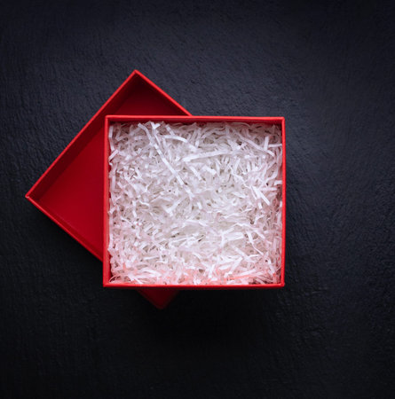 Top view of red box for gift filled with decorative shredded white paper on black background