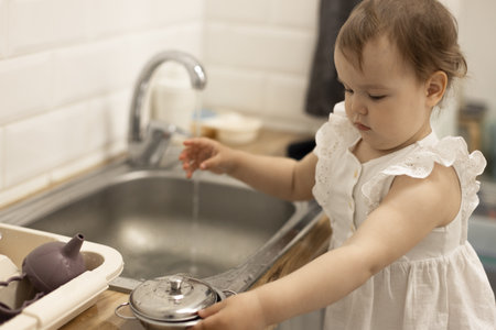 daughter 18-24 months plays with water and dishes in the sink, girl washes toy dishes, mom's helperの写真素材