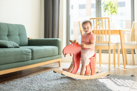funny baby girl sits in children's wooden rocking chair, child in rocking horse in living roomの写真素材