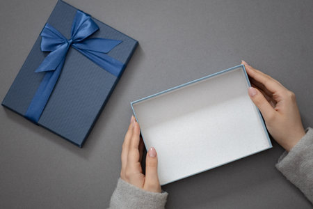 Open gift box in woman hands on gray background. Blue cardboard box with bow for your product. Flat...