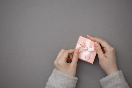 First person top view photo of hands unpacking craft paper gift box with pink satin ribbon bow over shiny sequins gray background with blank spaceの写真素材