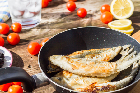 Fried fish in a frying pan, served with cherry tomatoes, lemon, olives and a glass of cold water with ice.の写真素材