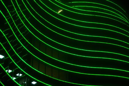 Green futuristic abstract wavy LED lines on building at night.  Abstract background from a stack of glowing LEDs in the fog. Bottom up view. Out of focus.の写真素材