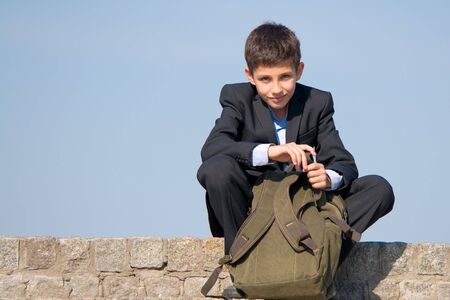 a successful school student is sitting on the stones and holding his schoolbag in his handsの写真素材