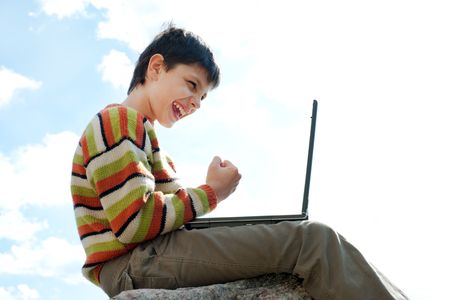 A shouting teen is sitting on the rock outside and playing a computer gameの写真素材