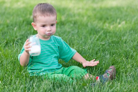 A serious little boy in green is drinking milk sitting on the green grassの写真素材