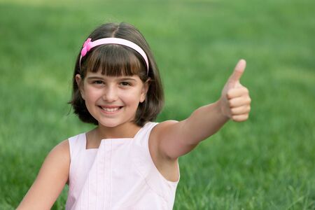 A pretty smiling girl in pink is sitting on the grass and holding her thumb upの写真素材