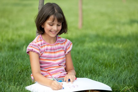 A portrait of a pretty smiling girl in pink sitting on the summer grass and drawingの写真素材