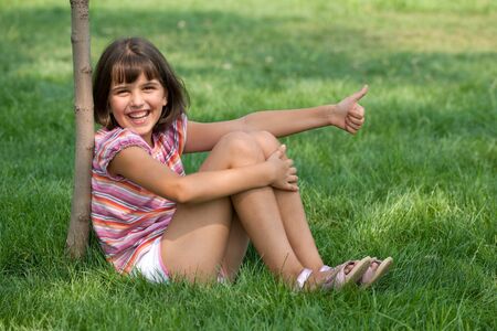 A pretty smiling girl is sitting on the grass and holding her thumb upの写真素材