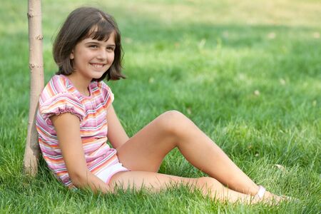 A pretty resting girl in pink is sitting on the summer grass by the young treeの写真素材