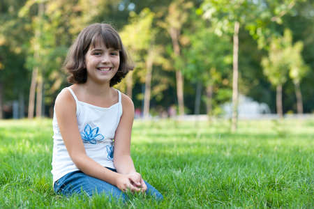 A portrait of a pretty smiling girl sitting on the green grass in the autumn parkの写真素材
