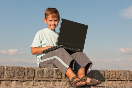 A handsome smiling boy is sitting outdoor with a laptop on his kneesの写真素材