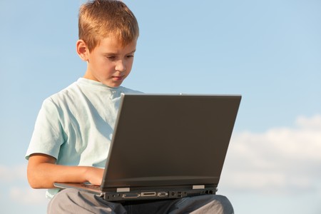 A boy with laptop is sitting outdoor against the skyの写真素材