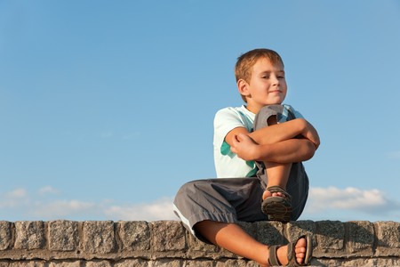 A handsome boy in blue is sitting outdoor against the sky hugging his kneeの写真素材