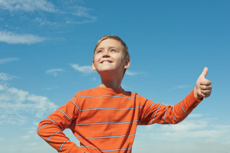A happy boy standing against the sky is showing his thumb up