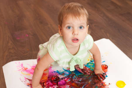 A toddler is painting a picture with her hands and legs on the big white sheet of paper lying on the wooden floorの写真素材