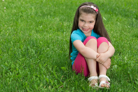 A cheerful little girl is sitting on the green grass in summerの写真素材