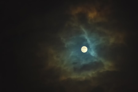 Full moon shines through dramatic sky surrounded cloudsの写真素材