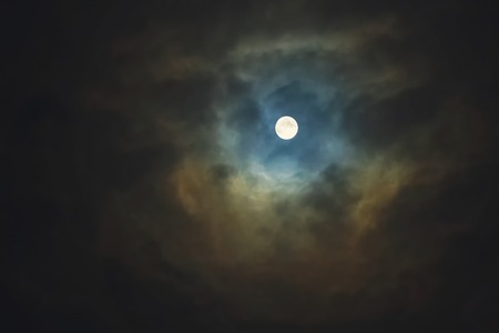 Full moon shines through dramatic sky surrounded cloudsの写真素材