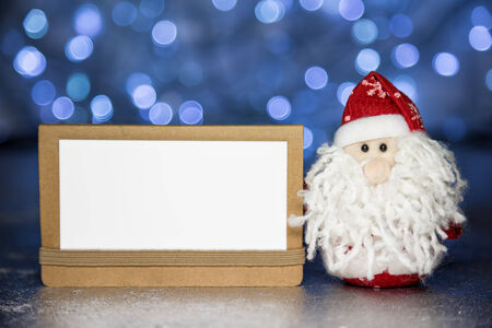 Santa Claus or Father Frost and greeting white blank card on craft paper with Christmas lights on backgroundの写真素材