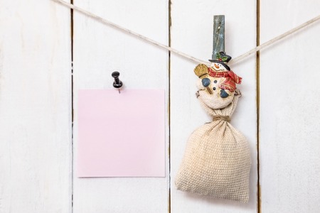Christmas snowman clothespin hanging on clothesline or rope and holding sack and pink blank note paper with push black pin on wood backgroundの写真素材