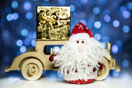 Santa Claus and old vintage wooden automobile with gift box on christmas lights background. Main focus of image on Santa Claus and defocused on side turned wood car with gift boxの写真素材