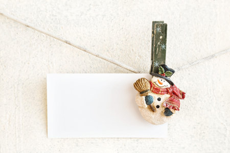 Christmas snowman clothespins hanging on clothesline or rope and holding white blank paper card on plaster backgroundの写真素材