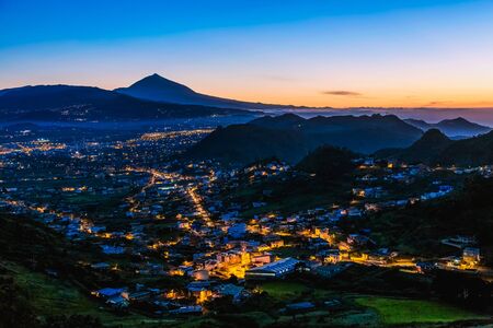 City or town with illumination after sunset or sundown at evening in mountains with blue sky and Teide volcano on background in Tenerife Canary island, Spainの写真素材