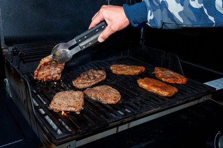 Hand flips a piece of juicy barbecue meat. The gas grill is black, it cooks delicious meat.
